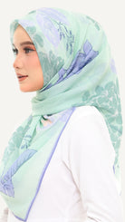 A woman wearing a mint green hijab with a light purple floral pattern, styled over a white top against a plain white background.