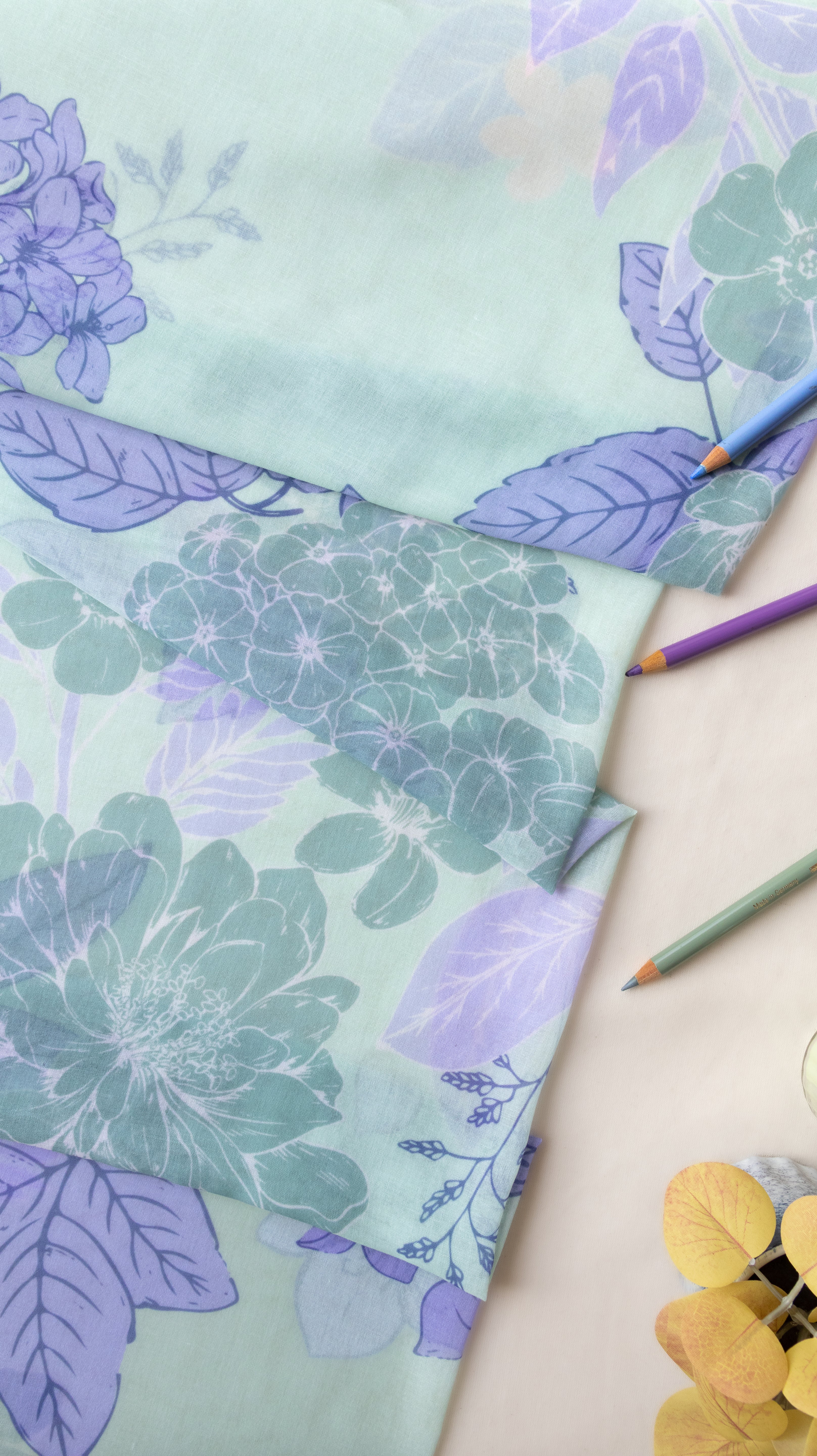 A close-up of patterned fabric with large floral and leaf motifs in soft mint green, lavender, and teal, arranged on a light surface with colored pencils and a small yellow flower prop.