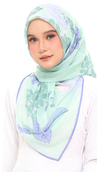A woman wearing a mint green hijab with a light purple floral pattern and a solid purple border, paired with a white long-sleeve top against a plain white background.