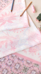 A styled flat-lay of light pink floral fabric with a delicate border, layered over a darker pink floral fabric. A spool of pink thread, two wooden pencils, and a small green leaf are arranged on top of the fabrics against a light beige background.