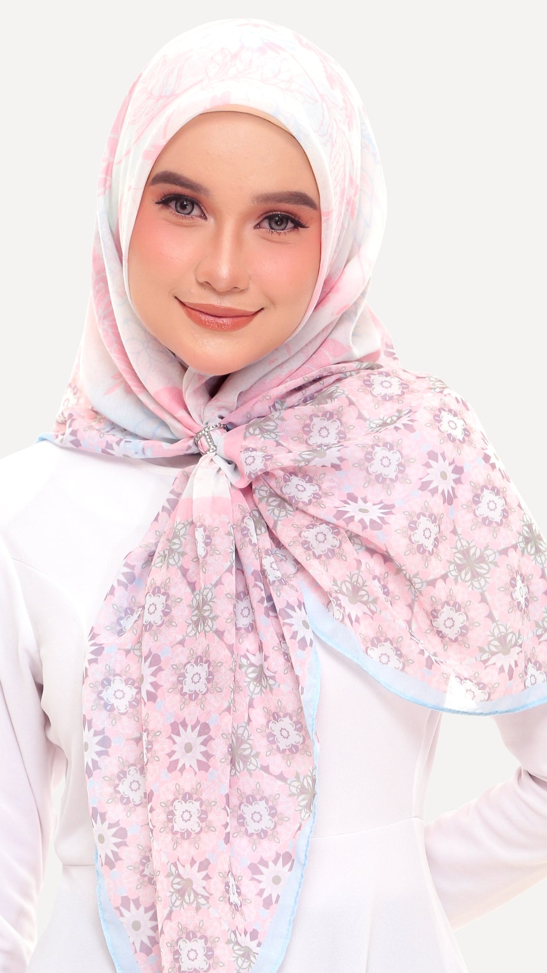 A woman wearing a light pink hijab with a white and gray floral pattern and a light blue border, styled over a white top against a light gray background.