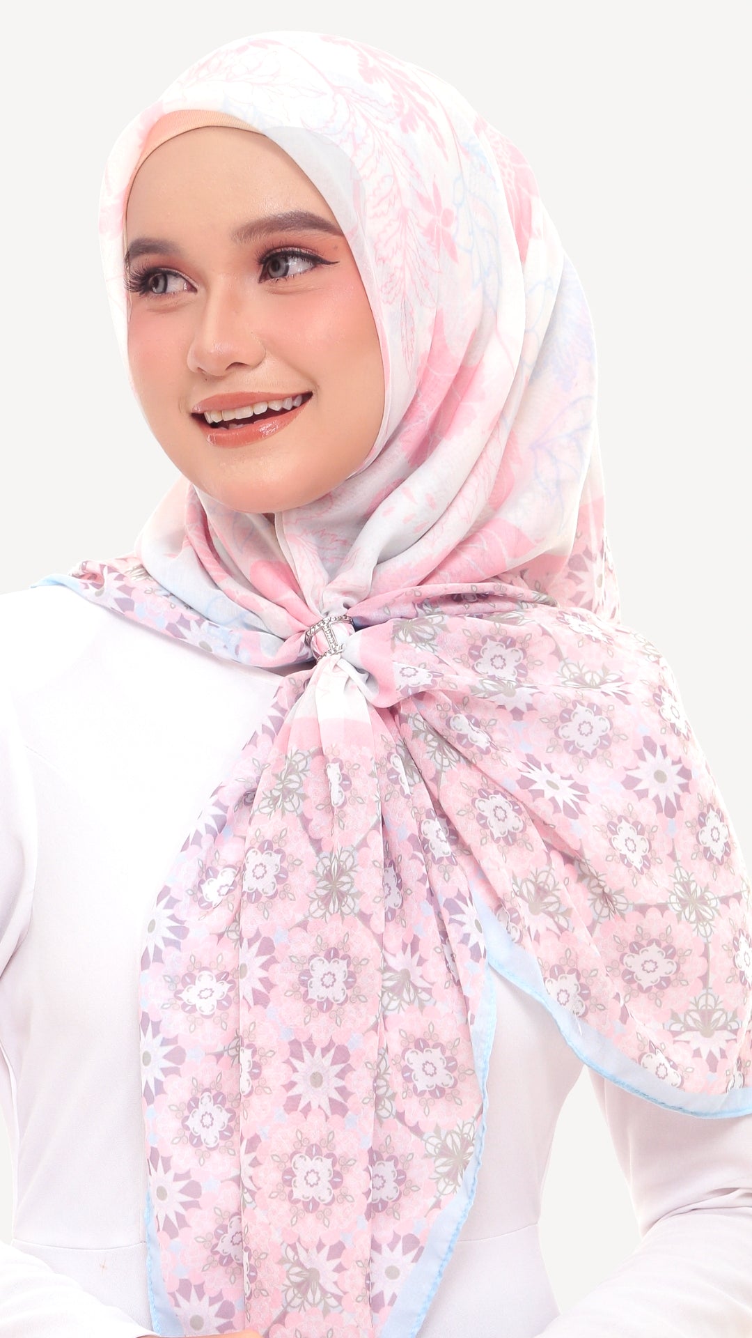 A woman wearing a light pink floral hijab with a white and pale blue border, tied neatly at the front. She is wearing a white long-sleeve top against a clean, light off-white background.