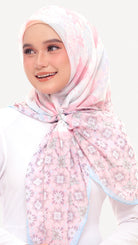 A woman wearing a light pink floral hijab with a white and pale blue border, tied neatly at the front. She is wearing a white long-sleeve top against a clean, light off-white background.