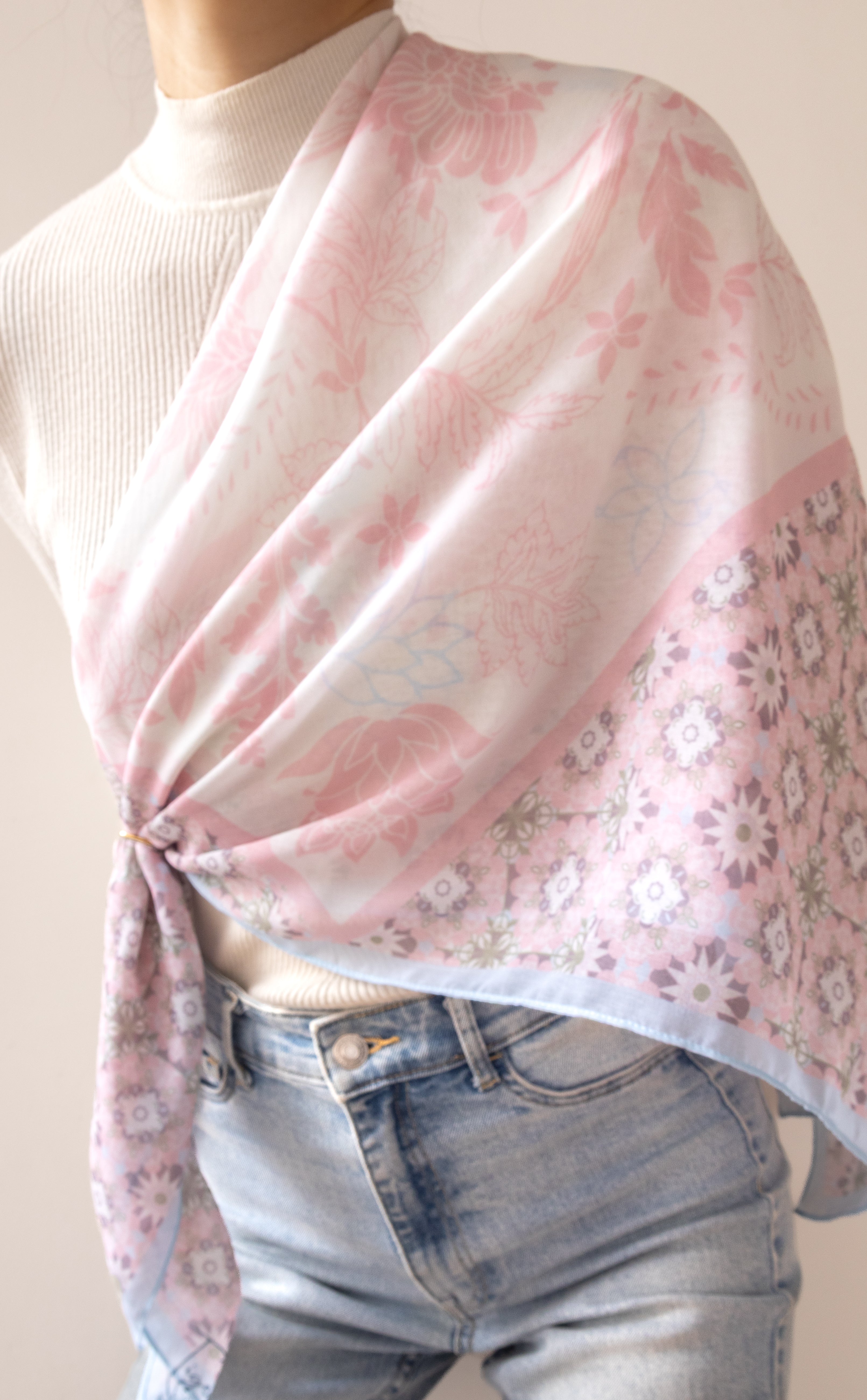 A light pink and white patterned scarf draped over a person's shoulder and tied at the waist, styled with a white ribbed turtleneck sweater and light-wash blue jeans against a plain beige background.