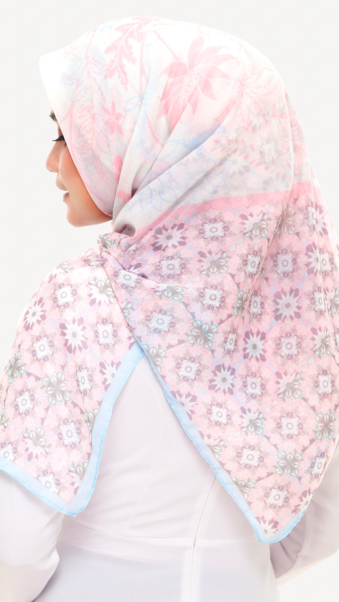 A woman wearing a light pink hijab with a floral pattern and light blue border, shown from a side-back angle against a plain white background.