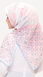 A woman wearing a light pink hijab with a floral pattern and light blue border, shown from a side-back angle against a plain white background.