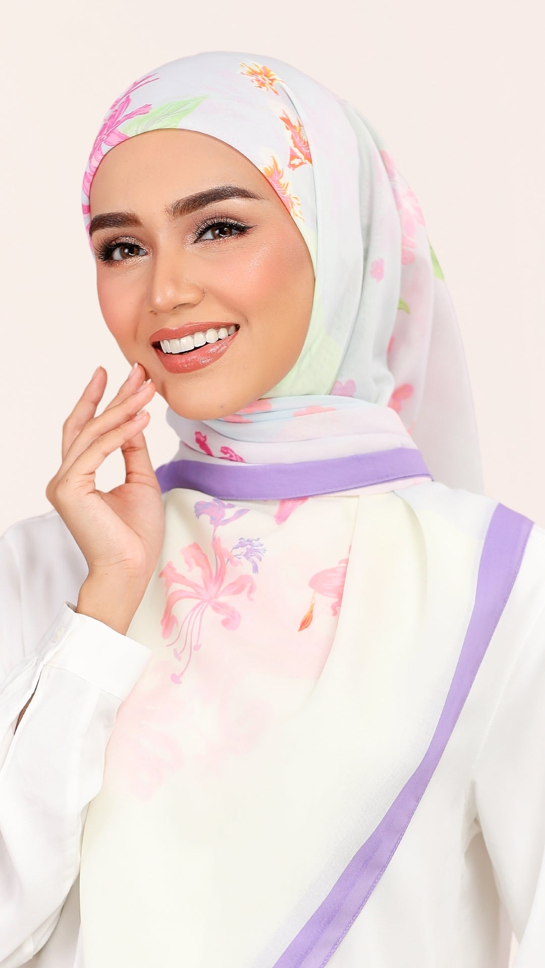 A woman wearing a white hijab with a pastel floral print in pink, purple, and green, paired with a white top and a purple headband, against a light beige background.