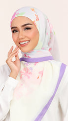 A woman wearing a white hijab with a pastel floral print in pink, purple, and green, paired with a white top and a purple headband, against a light beige background.