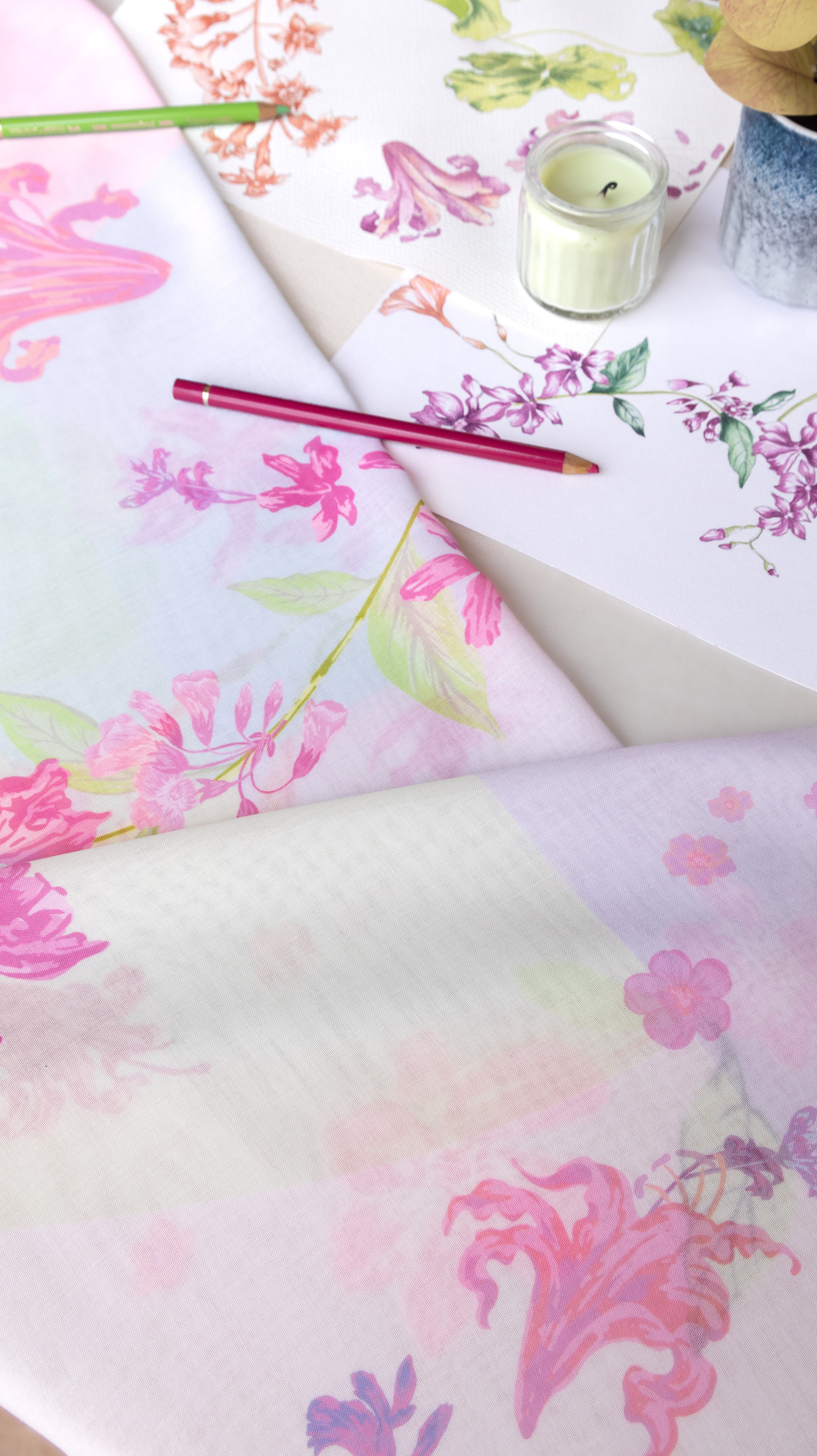 A styled flat-lay of two sheets of floral washi tape with pink, purple, and green botanical patterns on a white background, accented with a pink pencil and a small candle.