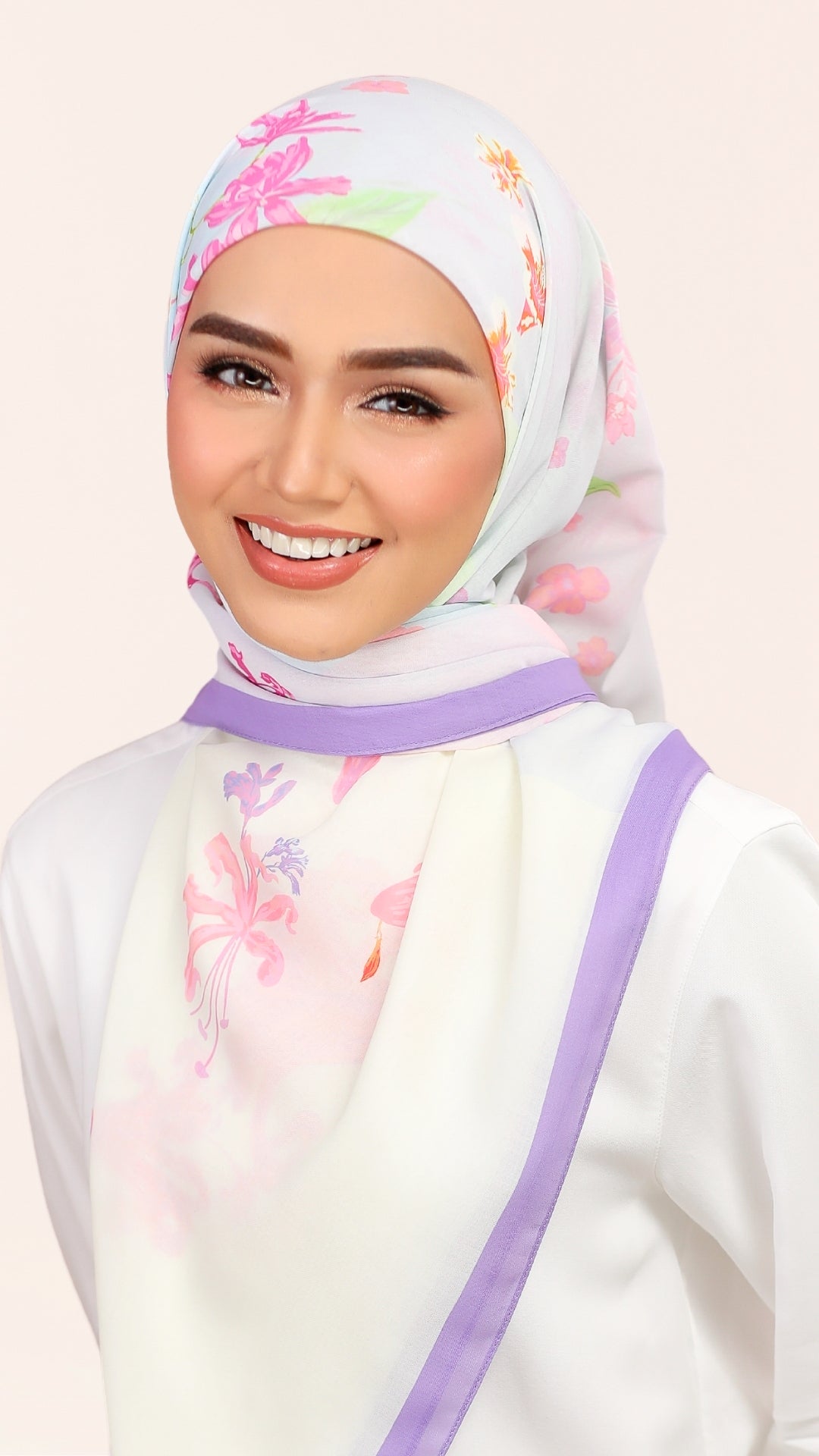 A smiling woman wearing a white hijab with pink and green floral patterns and a purple border, paired with a white top and purple shoulder strap, against a light beige background.