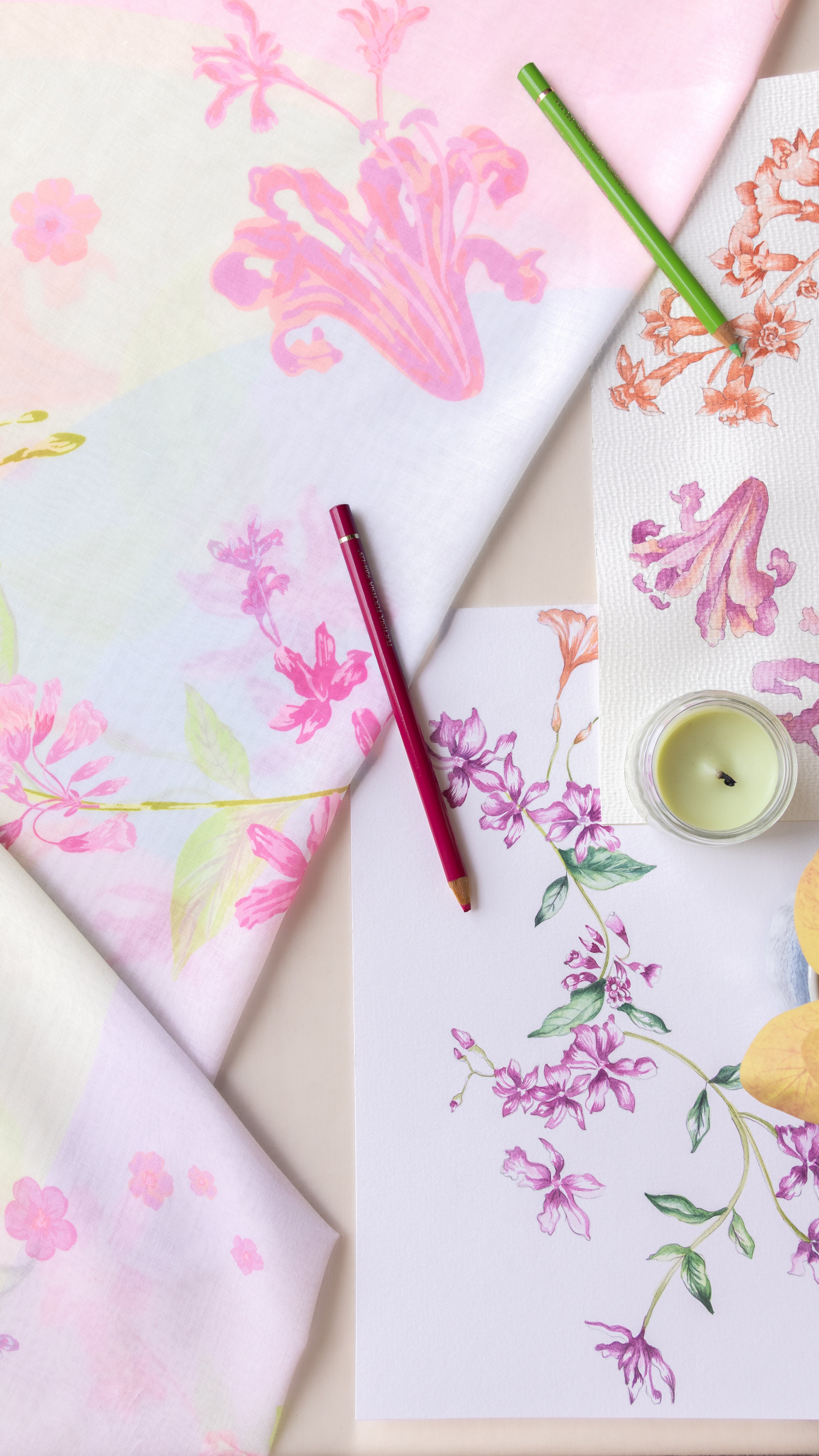 A styled flat-lay of floral printed textiles and paper sheets with pink, purple, and green botanical patterns, arranged with colored pencils and a small candle on a light neutral surface.