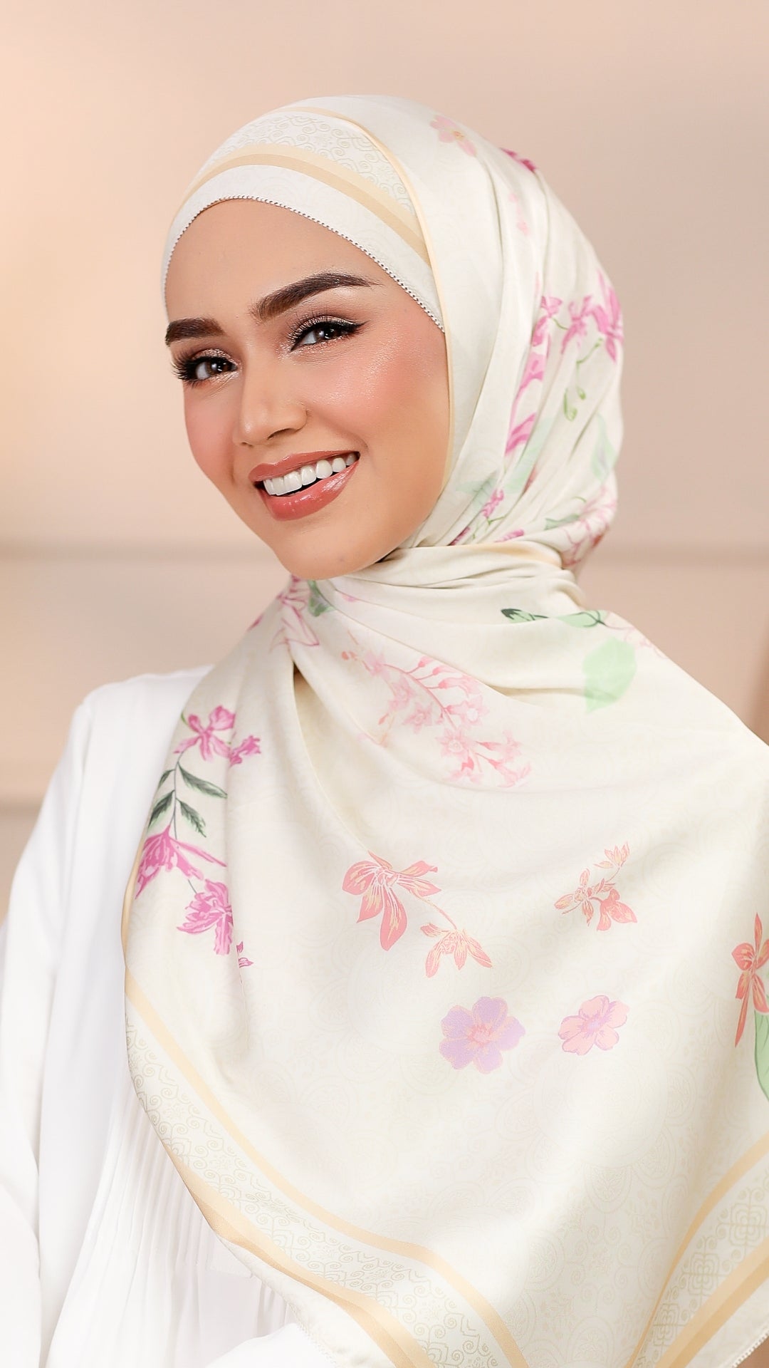 A smiling woman wearing a cream-colored hijab with a delicate pink and green floral pattern and gold border details, paired with a white top against a soft beige background.