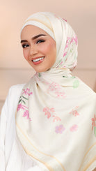 A smiling woman wearing a cream-colored hijab with a delicate pink and green floral pattern and gold border details, paired with a white top against a soft beige background.
