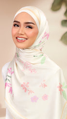 A smiling woman wearing a cream-colored hijab with a delicate pink and green floral pattern, photographed against a soft beige background with blurred green leaves.