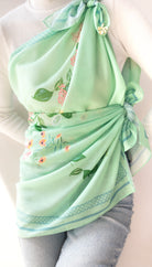 A mint green silk scarf with delicate floral and leaf patterns, styled as a halter top and wrap skirt on a model wearing a white ribbed sweater and light blue jeans.