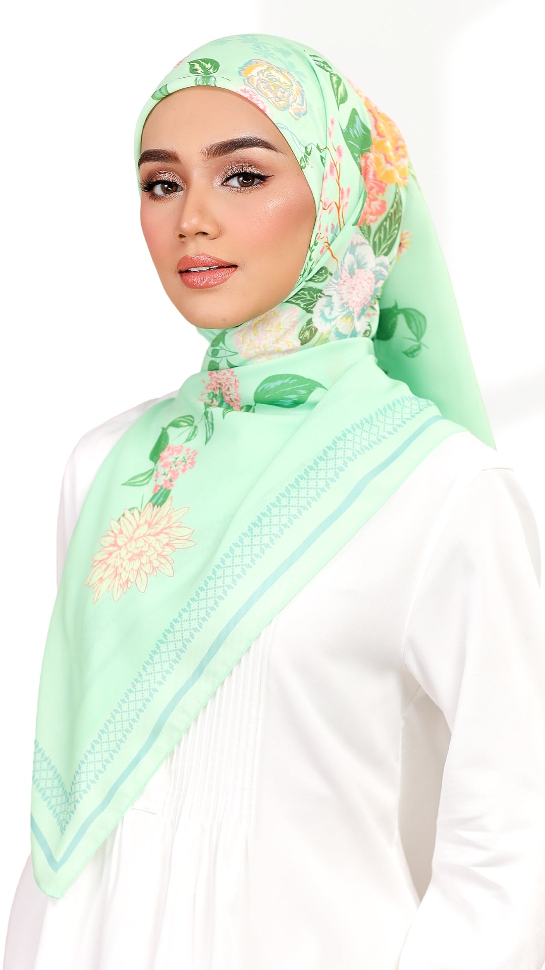 A woman wearing a mint green floral hijab with embroidered details and a white long-sleeve top, photographed against a clean white background.