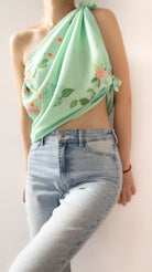 A mint green halter neck scarf with a floral and leaf pattern, worn by a model in light-wash blue jeans against a plain light background.