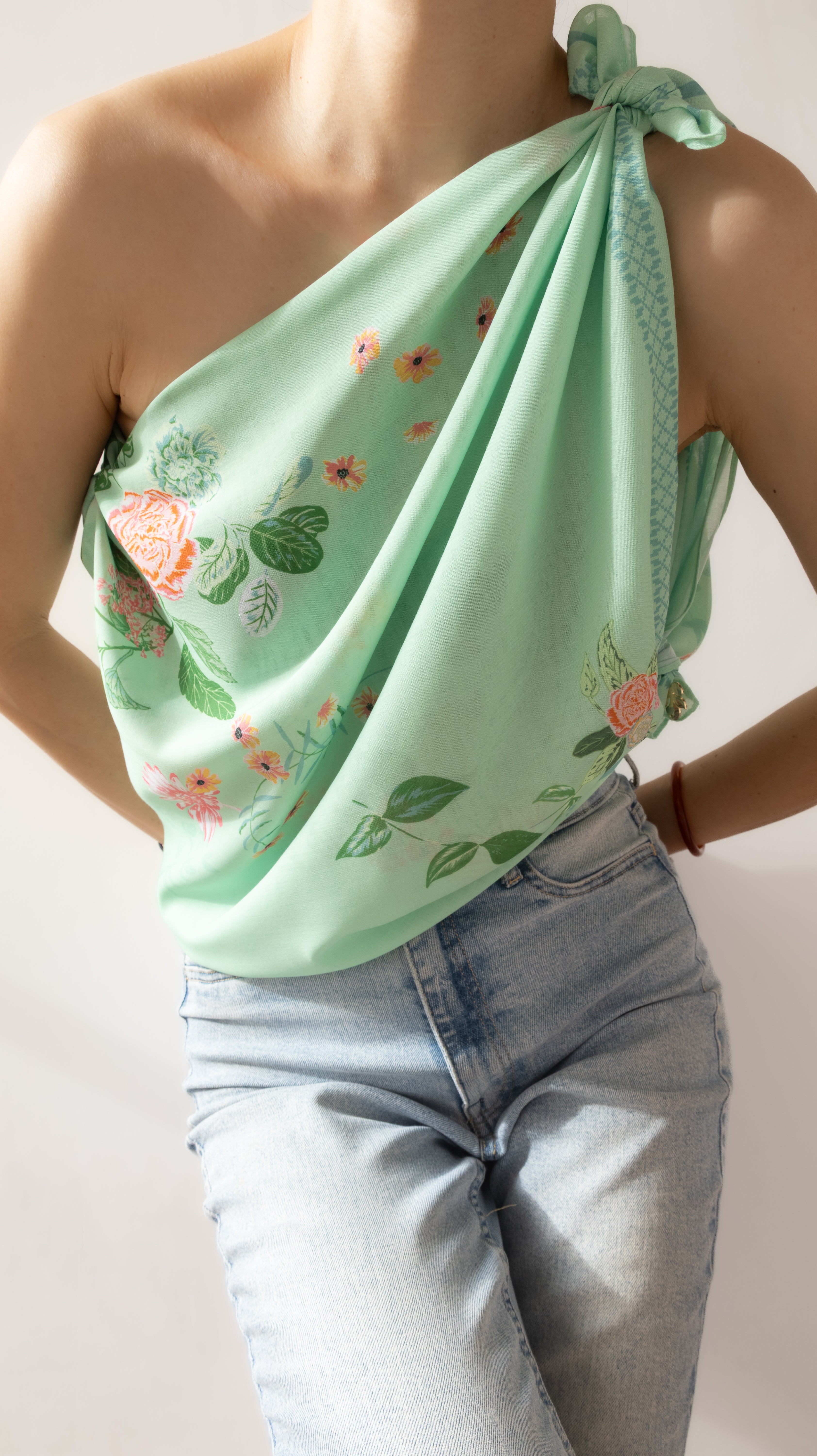 A mint green one-shoulder scarf top with a floral print, worn by a model in light-wash blue jeans against a plain off-white background.