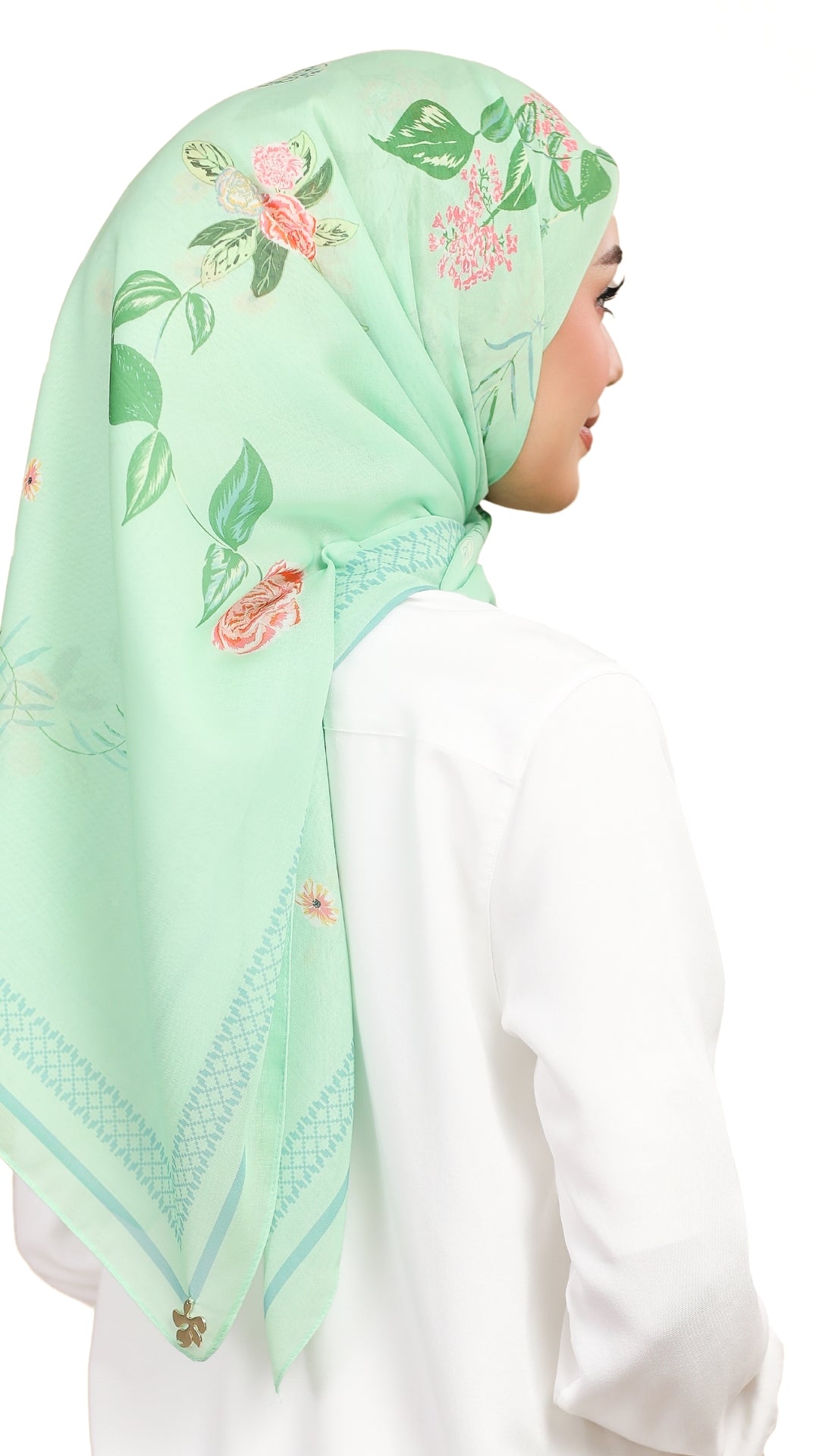 A woman shown from the back wearing a mint green hijab with pink floral prints and green leaves, paired with a white top against a plain white background.