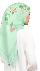 A woman shown from the back wearing a mint green hijab with pink floral prints and green leaves, paired with a white top against a plain white background.
