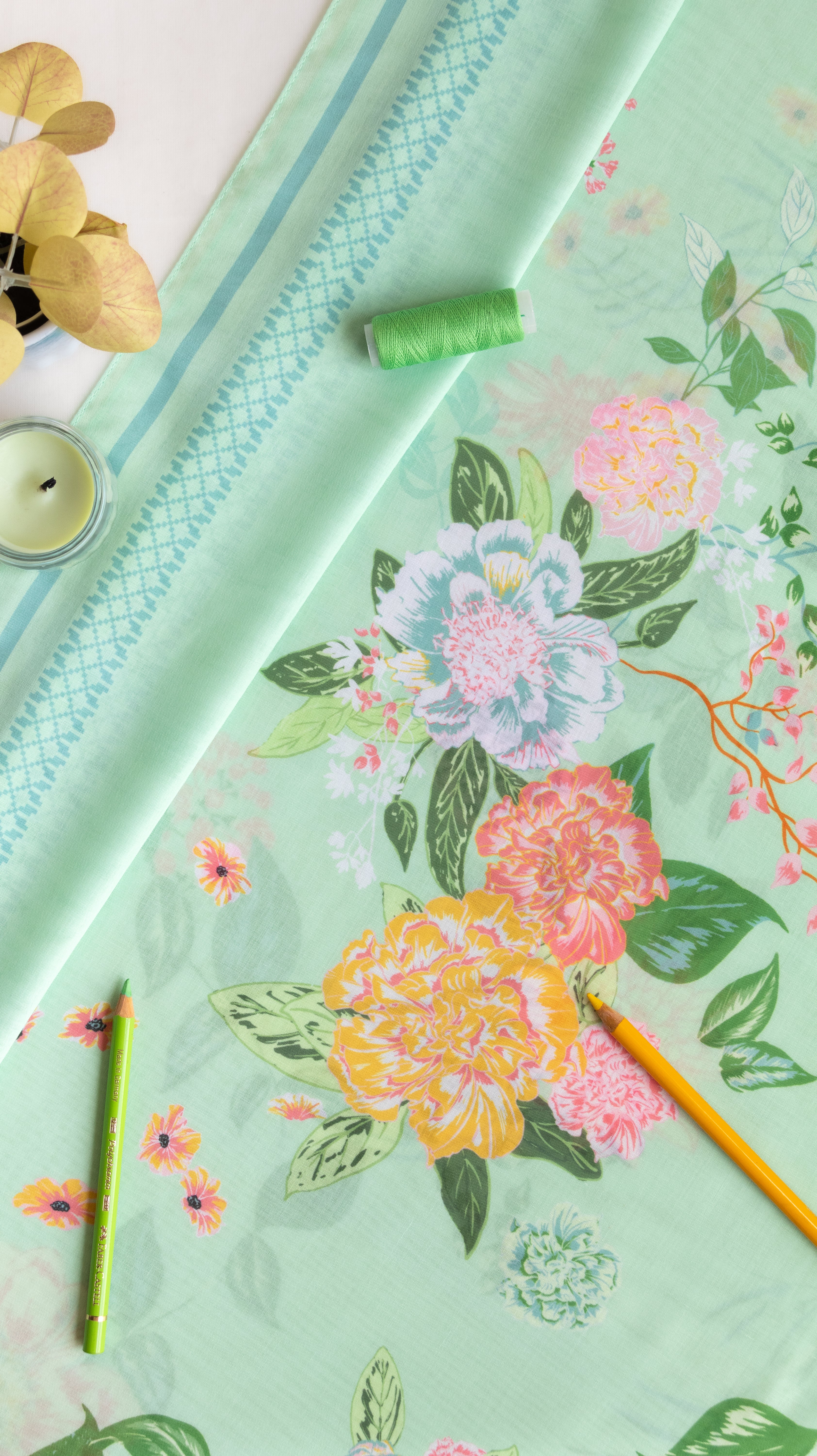 A styled flat-lay of a mint green fabric or wallpaper featuring a colorful floral print with peonies and leaves. The fabric is draped alongside a coordinating patterned ribbon, a green spool of thread, a small candle, and two colored pencils on a white surface.