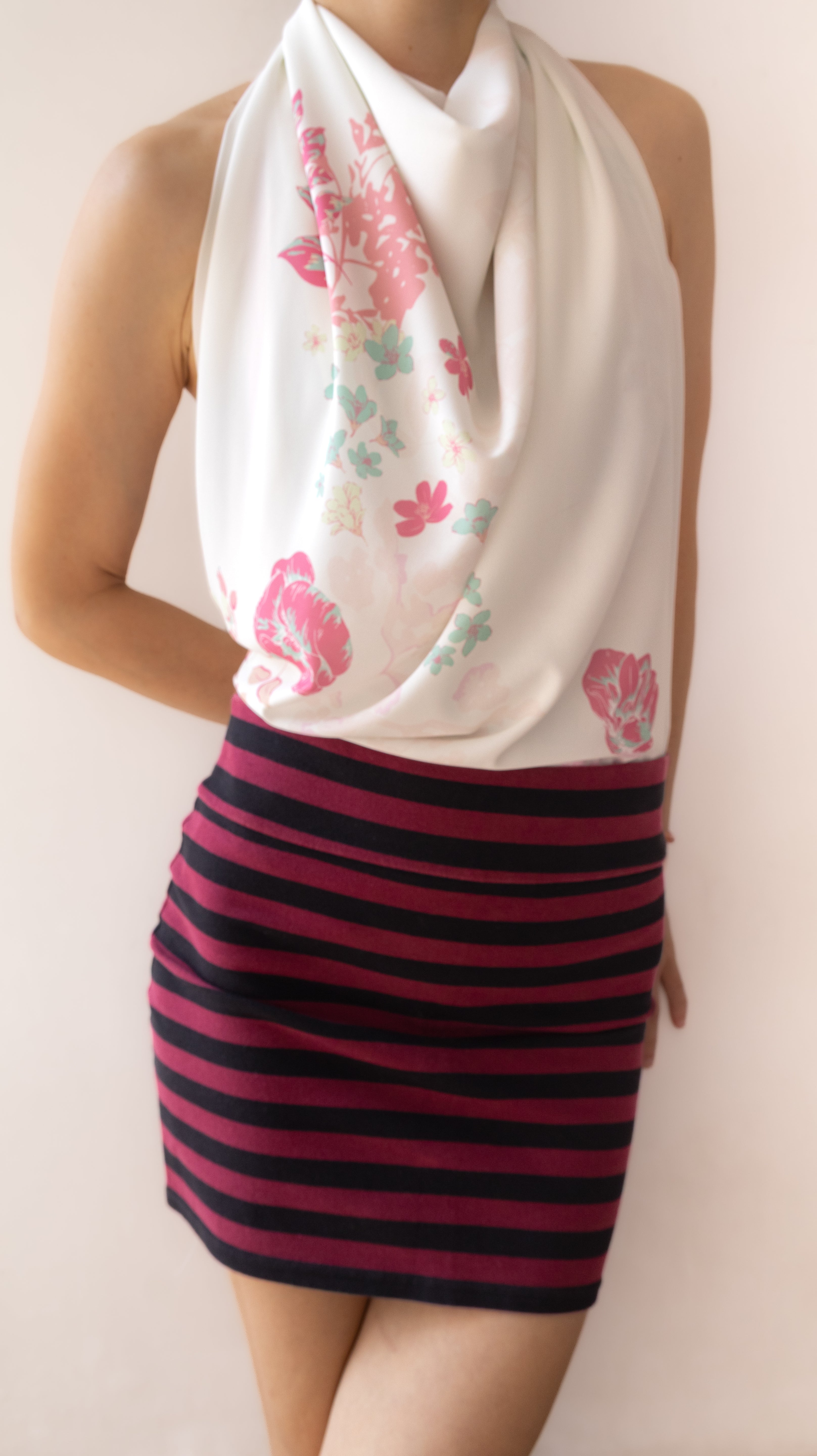 A model wearing a white halter-neck scarf with pink and green floral patterns, paired with a dark pink and black horizontally striped mini skirt, set against a plain beige background.