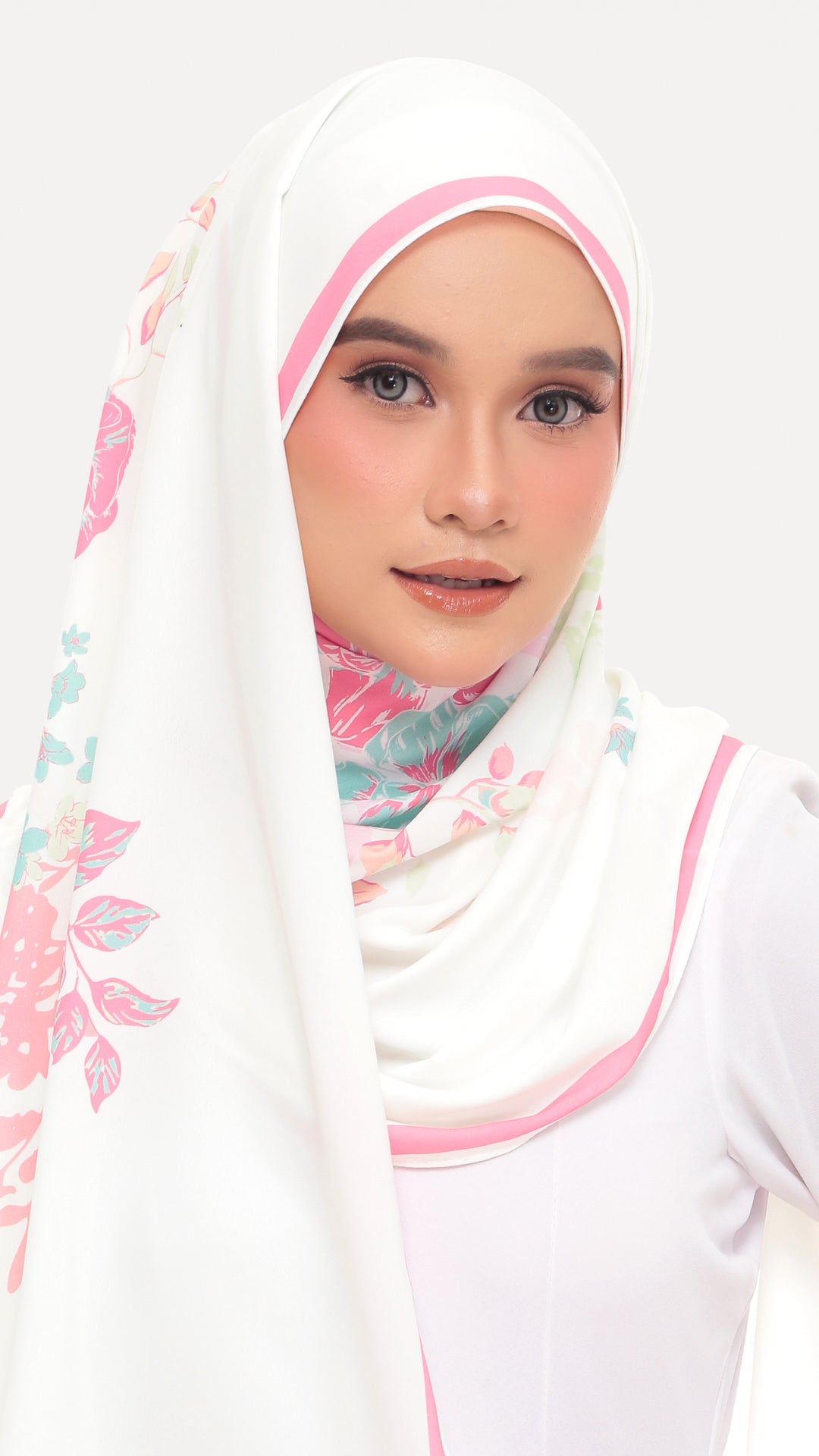 A woman wearing a white hijab with pink trim and a pastel floral print, paired with a white top, set against a plain light gray background.