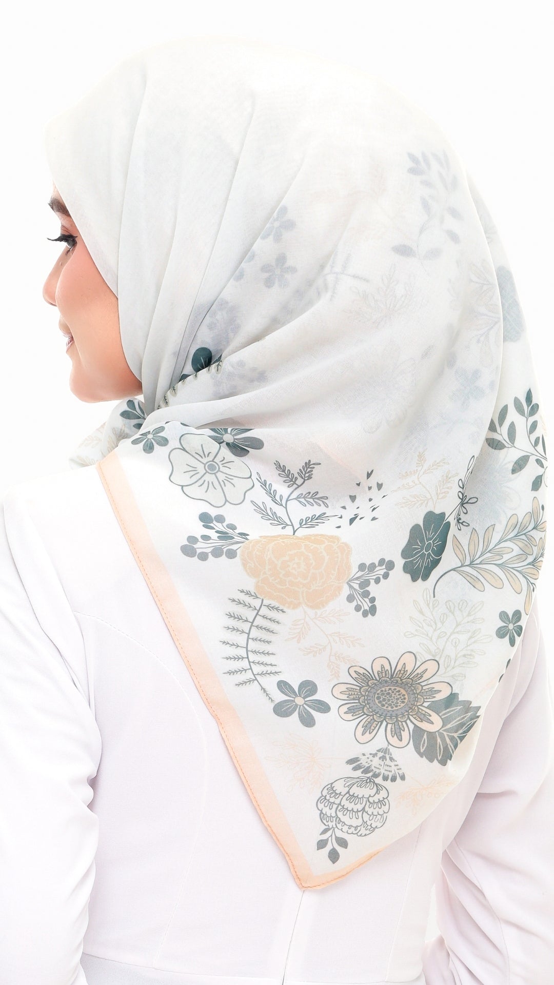 A woman wearing a white hijab with a delicate floral print in muted blue-gray and soft peach, shown from the side against a plain white background.