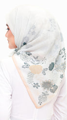 A woman wearing a white hijab with a delicate floral print in muted blue-gray and soft peach, shown from the side against a plain white background.