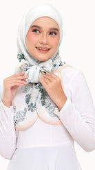 A woman wearing a white hijab with a green floral pattern and a peach-colored collar, styled with a white long-sleeve top against a plain white background.
