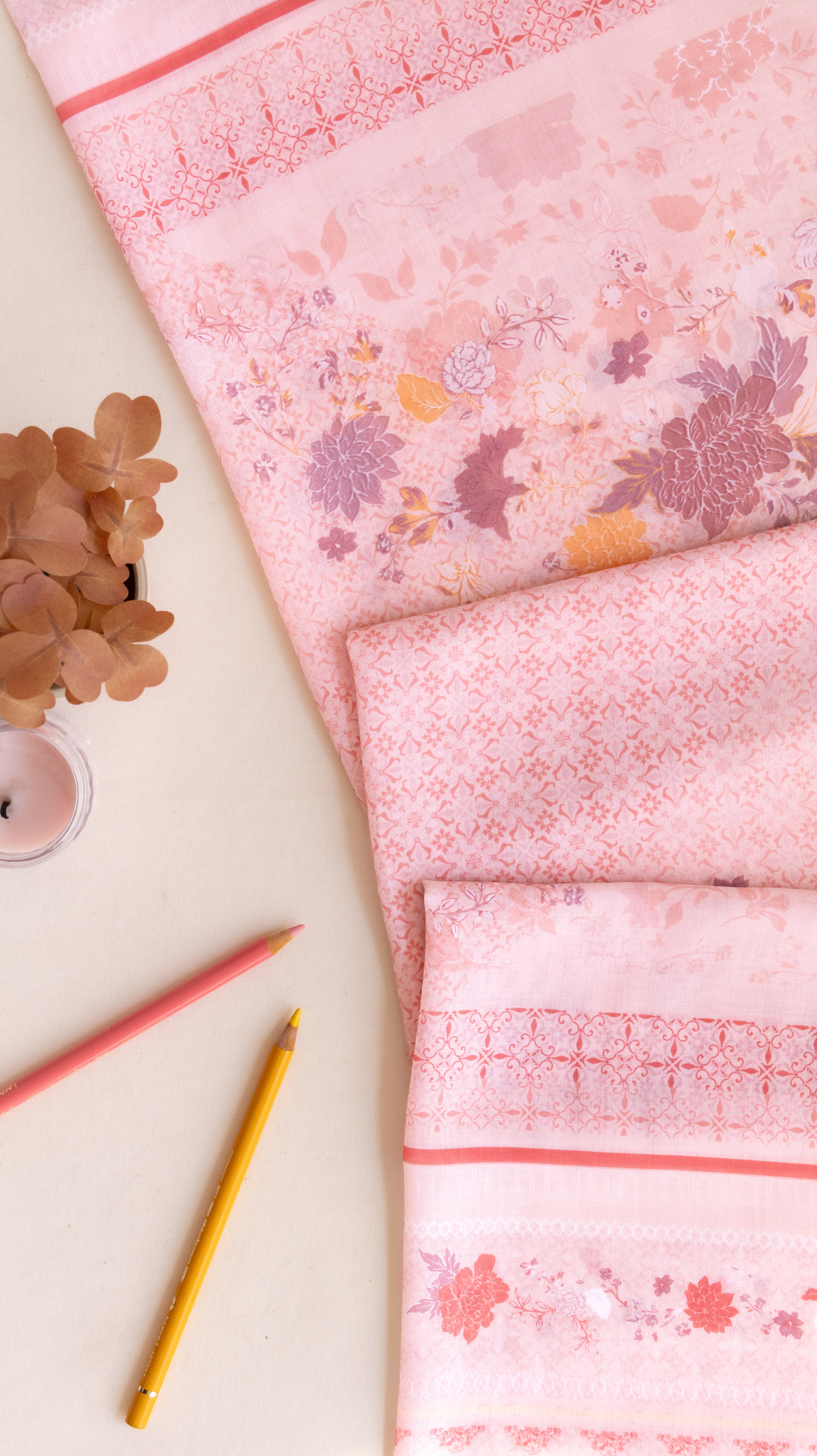 A pink patterned fabric, likely a kimono robe or sari, styled in a flat-lay arrangement on a light beige background with decorative dried flowers, a small candle, and two colored pencils.
