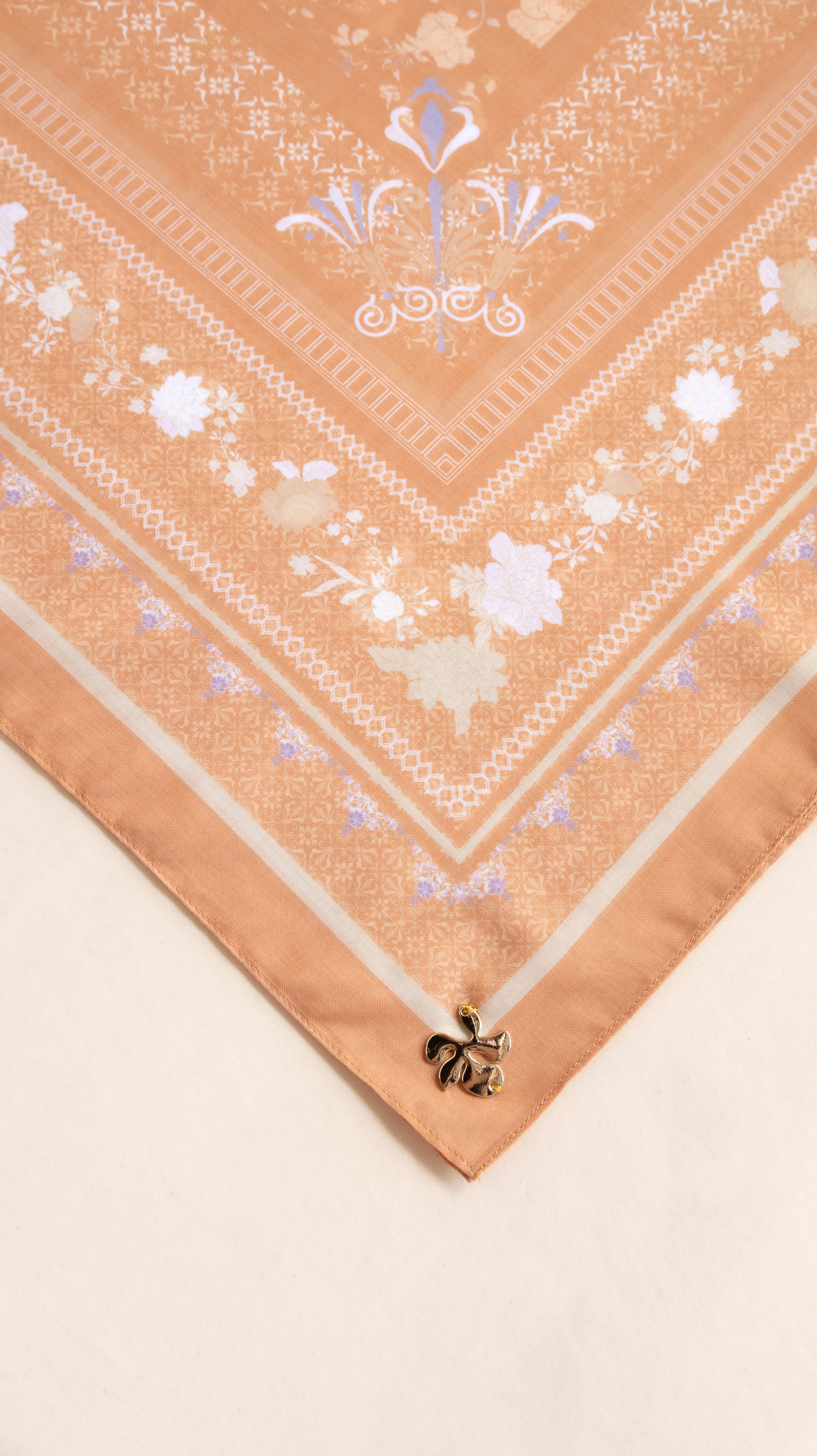 A close-up of a peach-colored silk scarf with white and light blue floral patterns and borders, laid diagonally on a light beige surface.