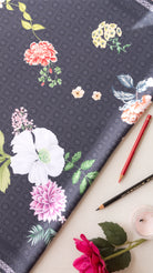 Dark gray floral fabric with white, pink, and yellow flowers, styled in a flat lay with colored pencils, a candle, and a rose.