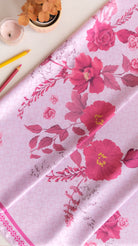A folded light pink fabric with a vibrant magenta floral print, styled on a light beige surface with decorative props including a small potted plant, a candle, and colored pencils.