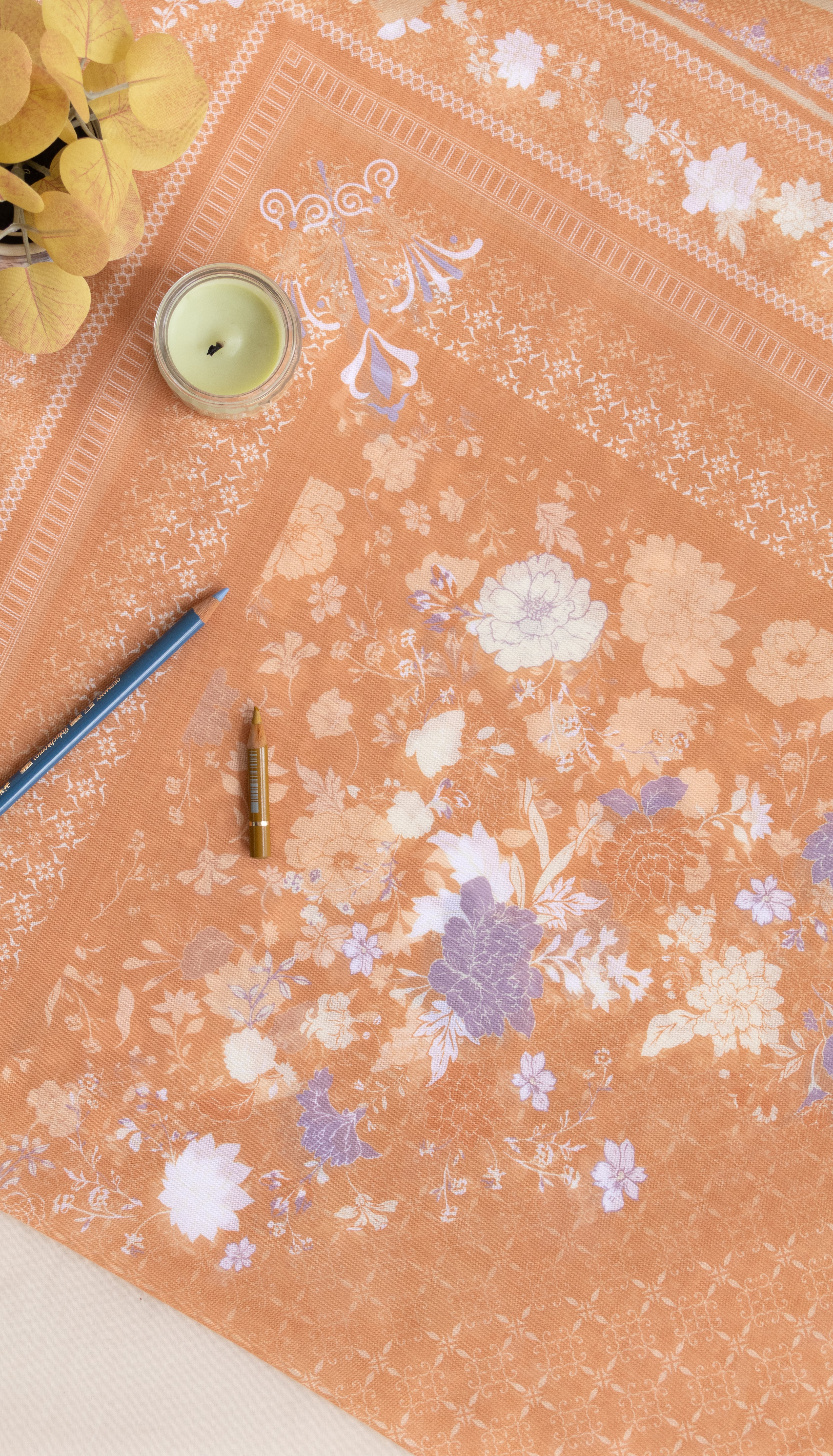 A peach-colored fabric or paper sheet with a delicate white and lavender floral pattern, laid out on a light surface with decorative props including a candle, pencil, and small bottle.