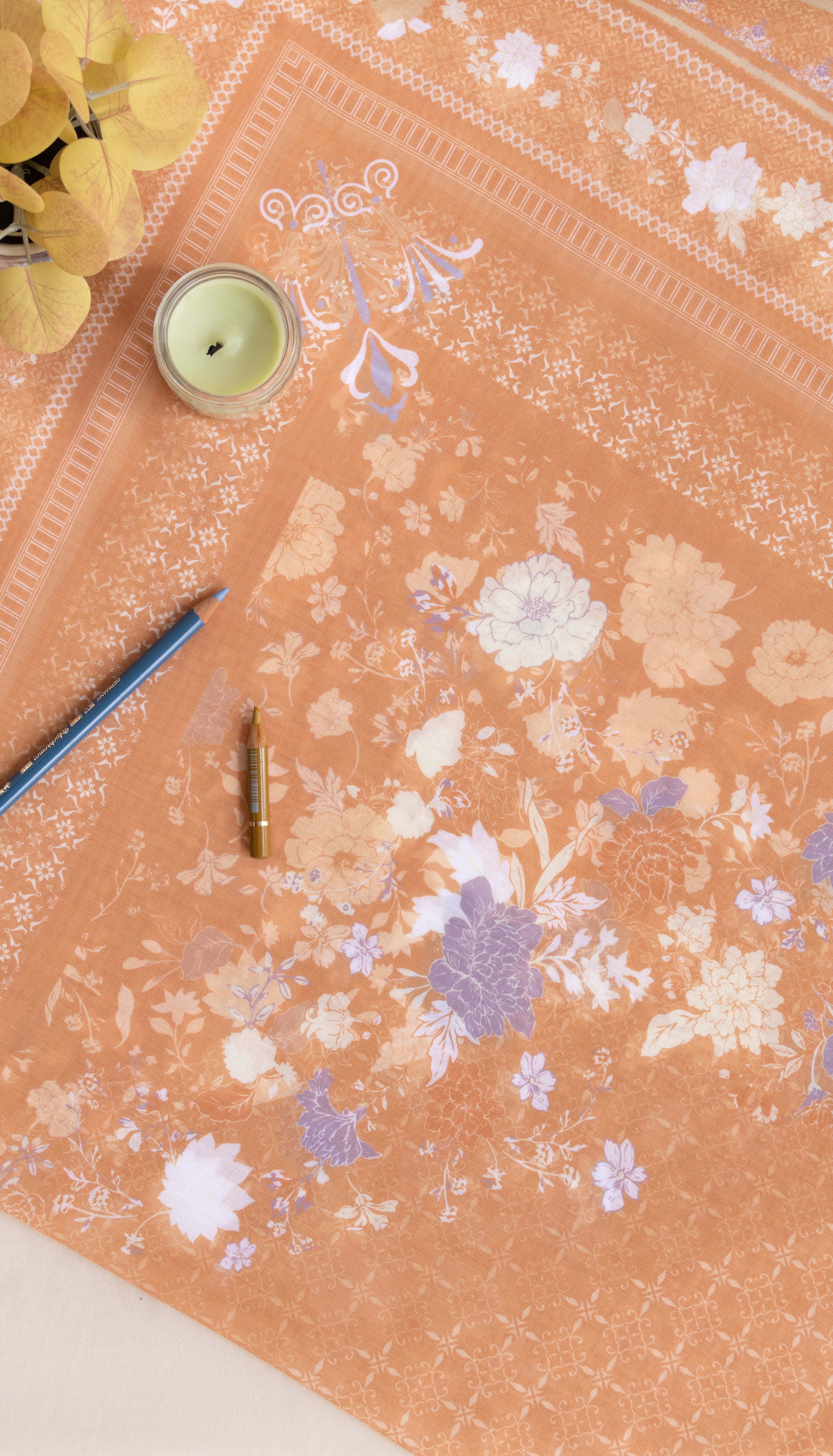 A peach-colored fabric or paper sheet with a delicate white and lavender floral pattern, laid out on a light surface with decorative props including a candle, pencil, and small bottle.