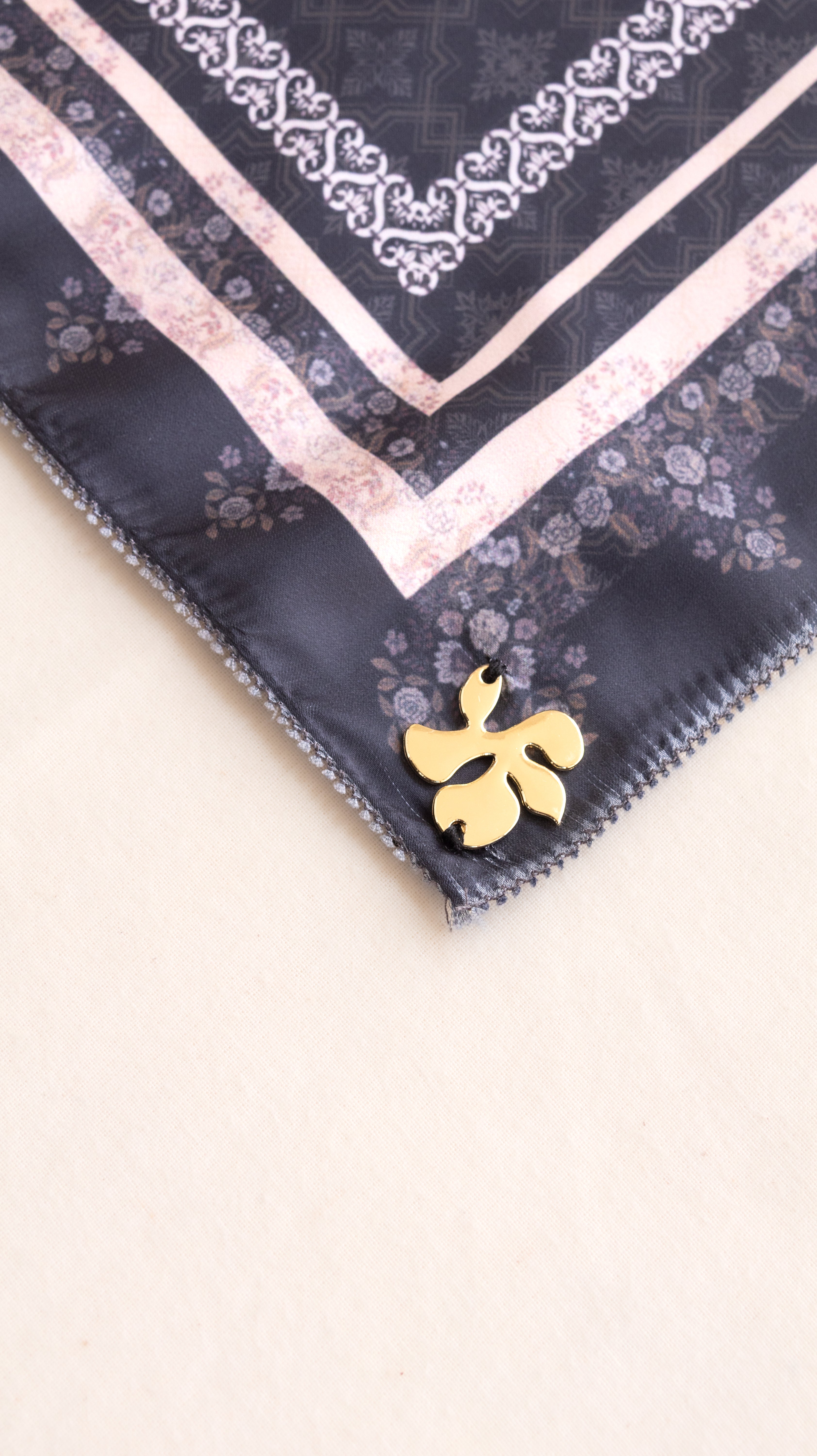 A close-up of a square scarf corner featuring a dark navy base with a light pink floral border, white ornamental trim, and a small gold flower-shaped pin attached near the edge, set against a plain light beige background.