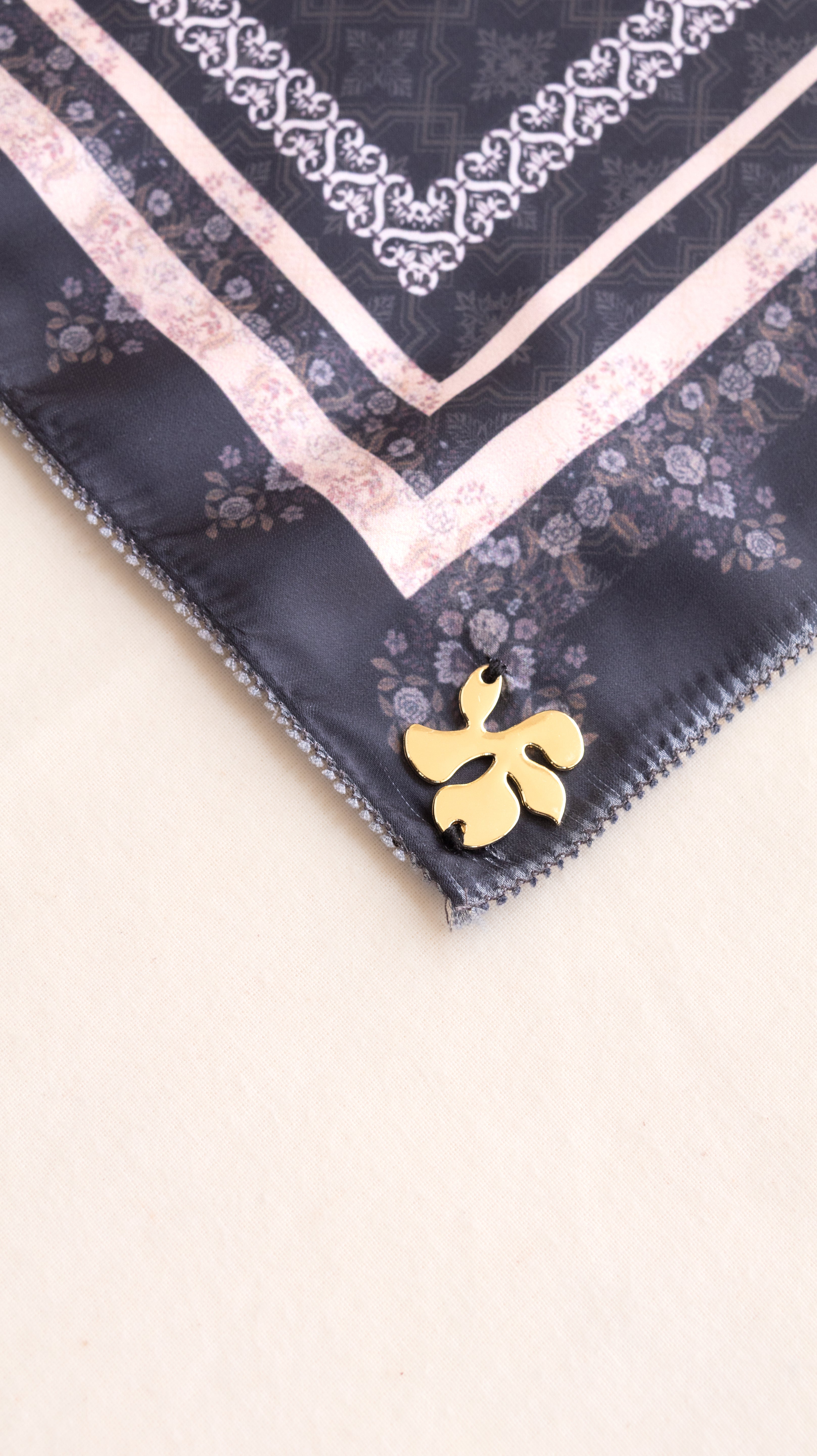 A close-up of a square scarf corner featuring a dark navy base with a light pink floral border, white ornamental trim, and a small gold flower-shaped pin attached near the edge, set against a plain light beige background.