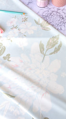 A close-up of a floral fabric or wallpaper sample with large pale pink and white flowers, muted green leaves, and a soft lavender background. A teal pencil and pink decorative items are partially visible at the top edge.