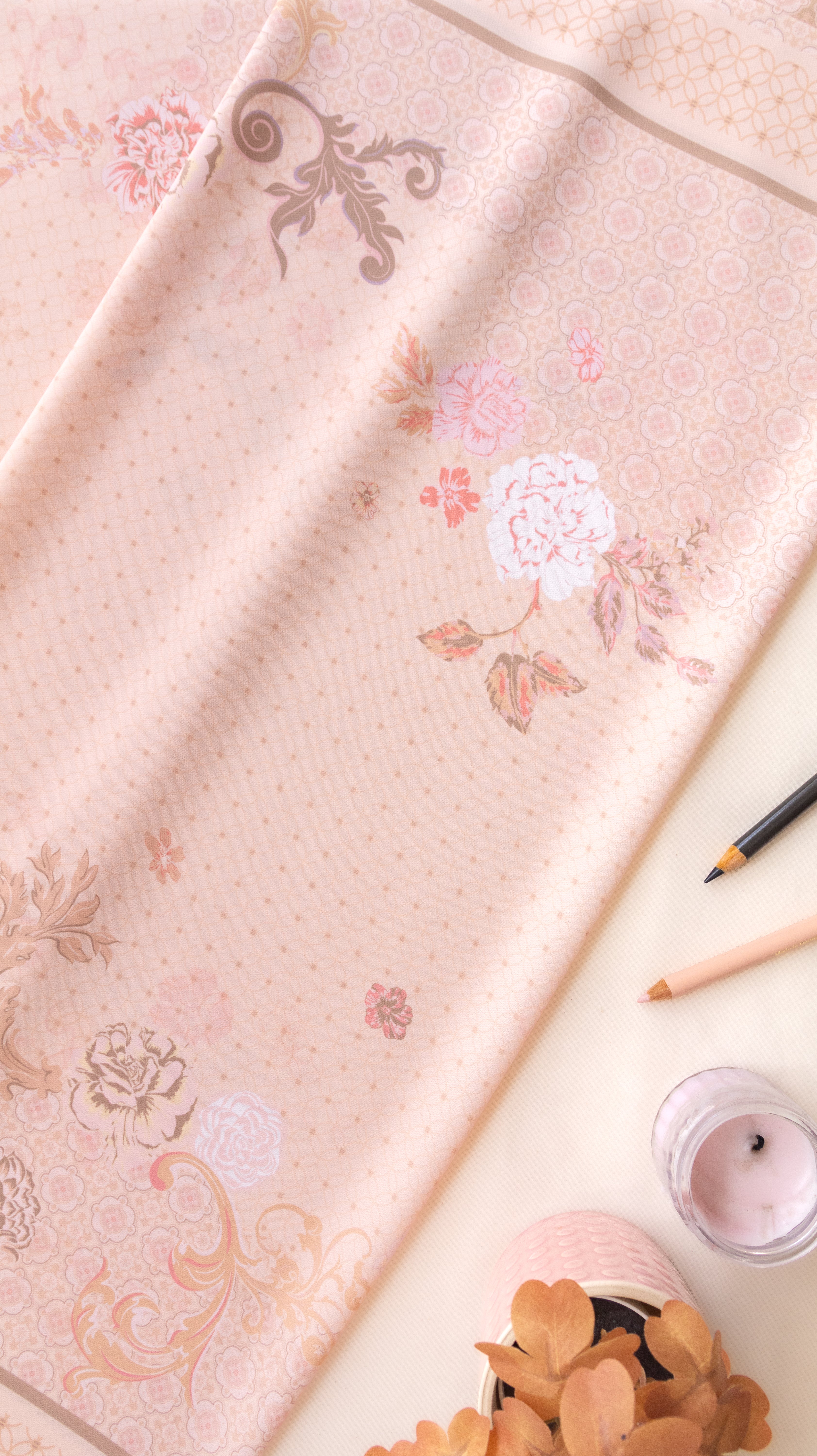 A light pink fabric with a delicate floral and scroll pattern, styled in a flat-lay composition with a pencil, a candle, and dried leaves on a cream background.