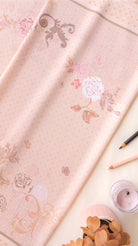 A light pink fabric with a delicate floral and scroll pattern, styled in a flat-lay composition with a pencil, a candle, and dried leaves on a cream background.