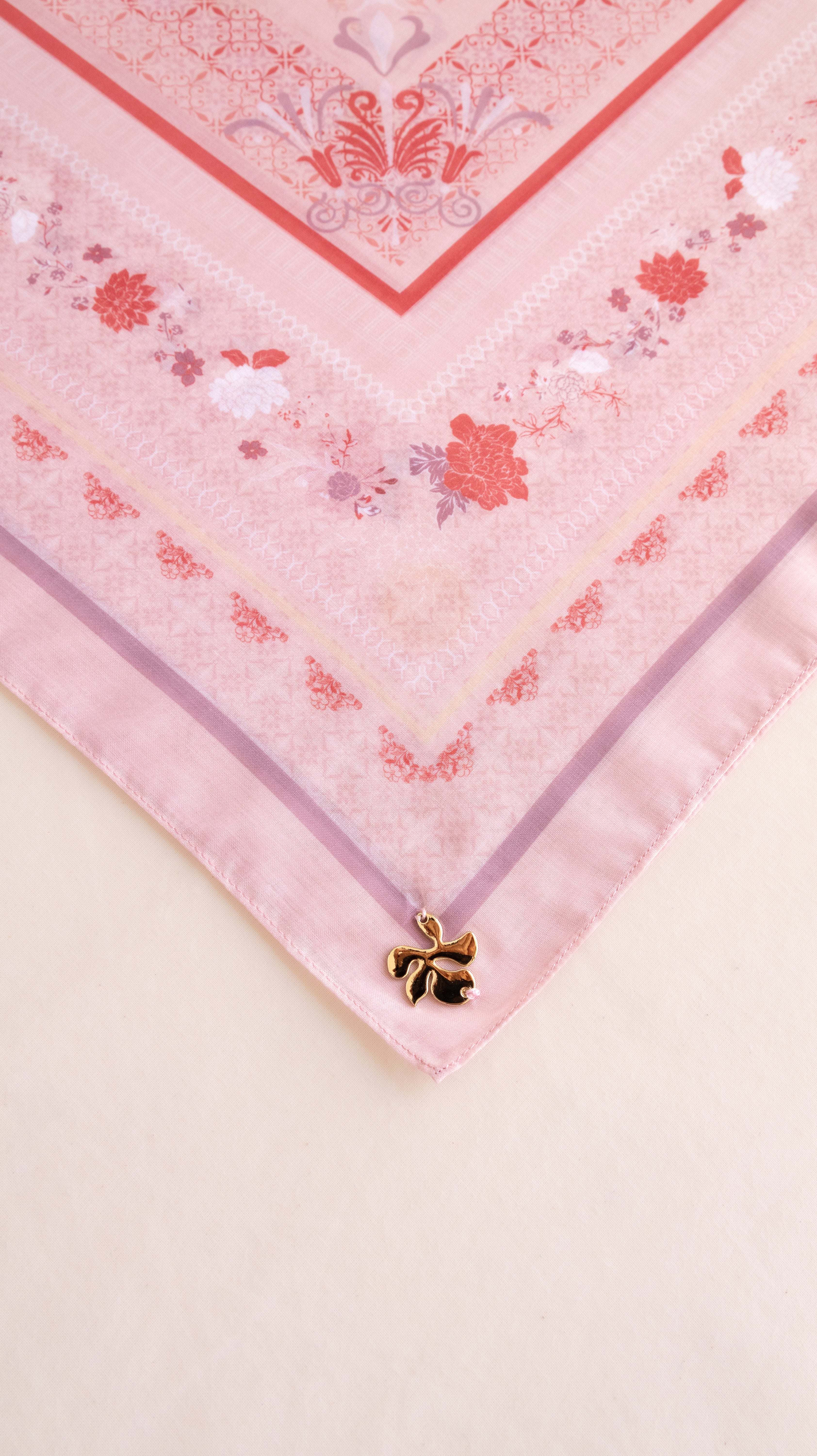 A close-up of a pink patterned scarf or bandana laid flat on a light beige surface, with a small gold bow charm attached near one corner.