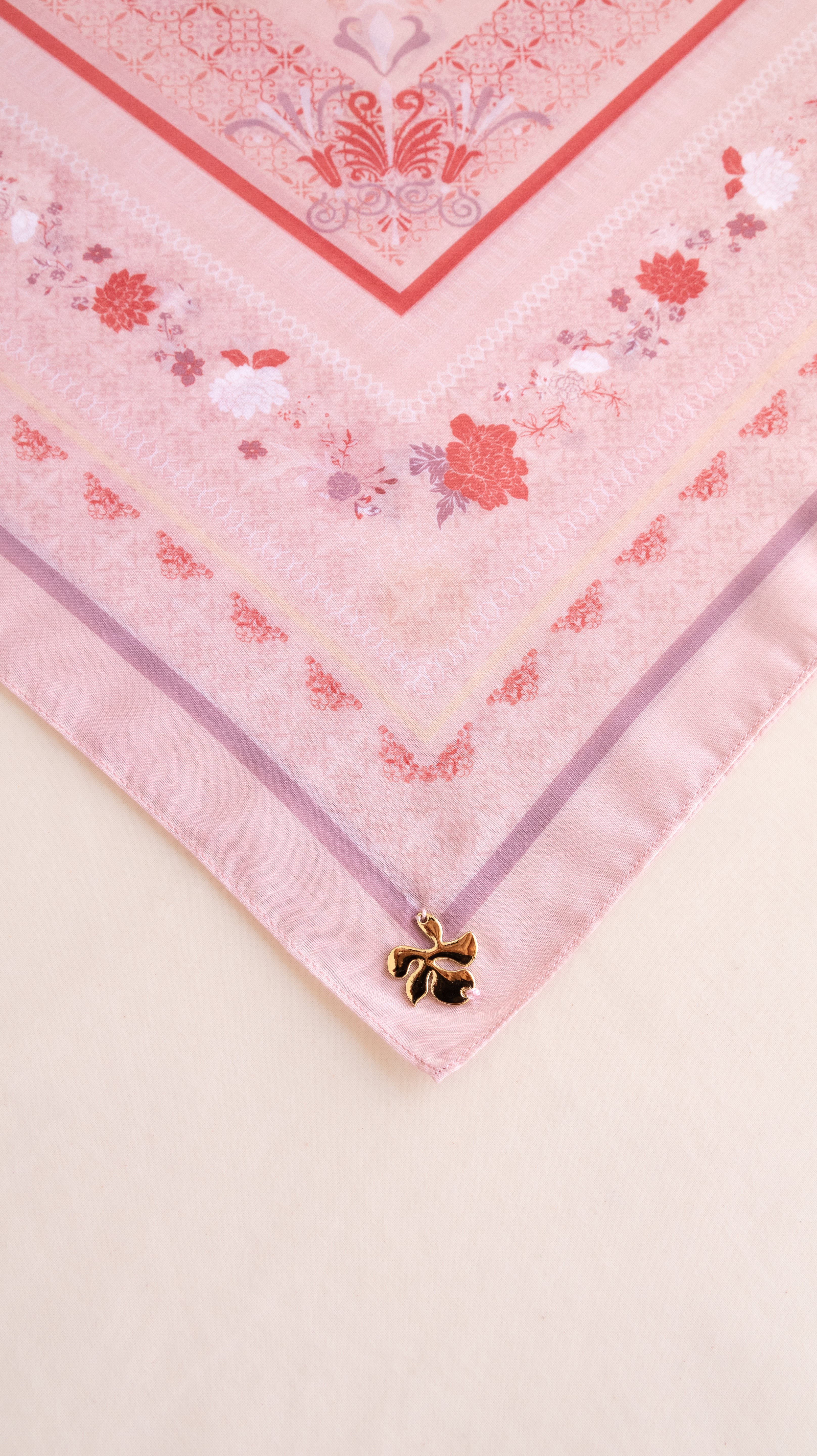 A close-up of a pink patterned scarf or bandana laid flat on a light beige surface, with a small gold bow charm attached near one corner.