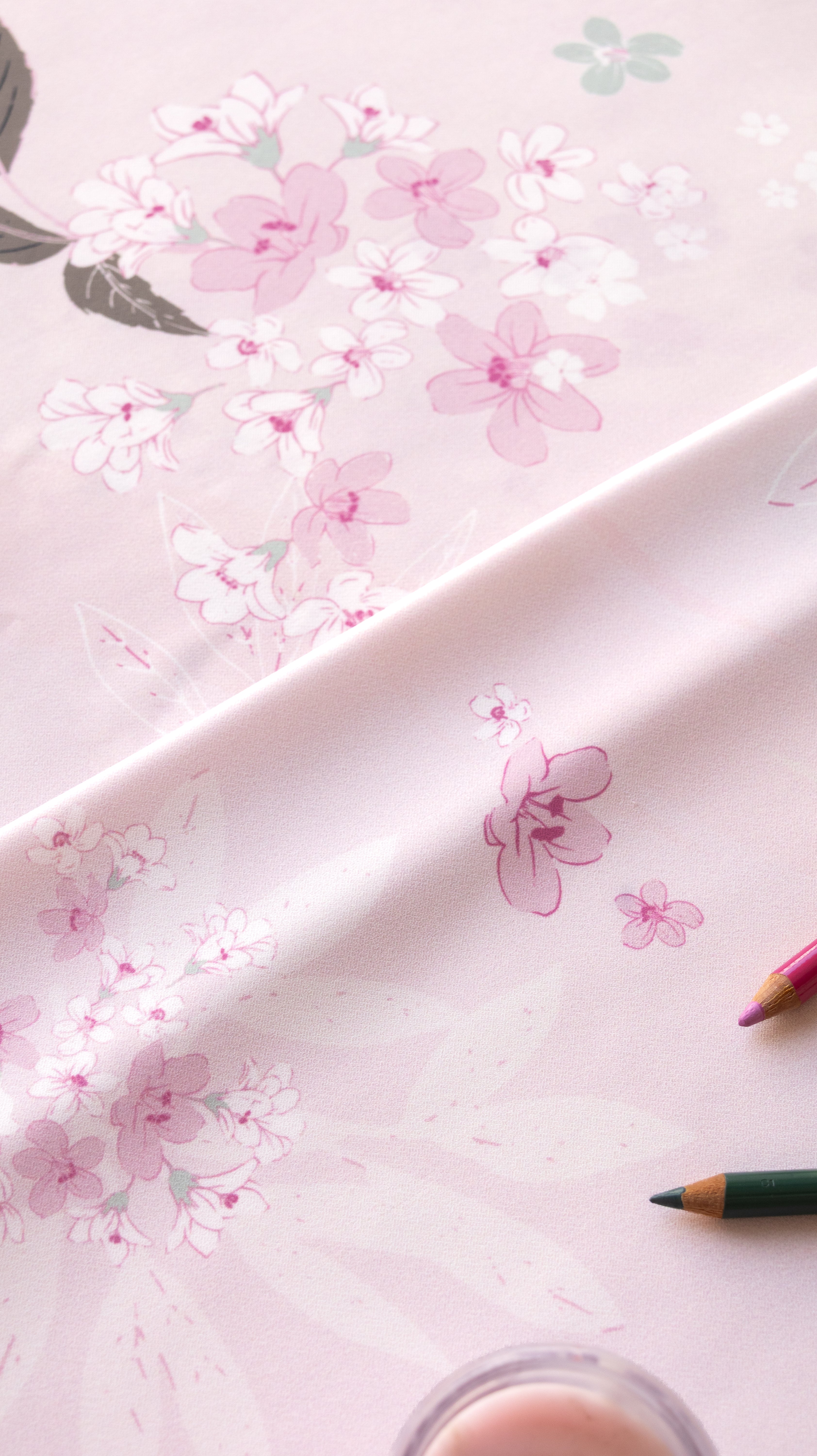 A close-up of a light pink fabric or paper with a delicate floral print in soft pink, white, and muted green, draped diagonally with colored pencils partially visible at the edges.