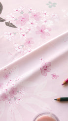 A close-up of a light pink fabric or paper with a delicate floral print in soft pink, white, and muted green, draped diagonally with colored pencils partially visible at the edges.
