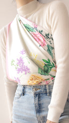 A close-up of a person wearing a cream ribbed mock-neck top with a floral printed scarf draped over the shoulder, paired with light-wash blue jeans.