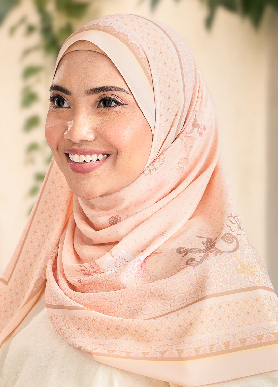 Woman wearing a peach hijab with intricate patterns, smiling outdoors.
