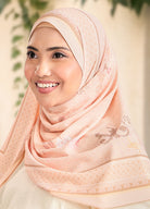 Woman wearing a peach hijab with intricate patterns, smiling outdoors.