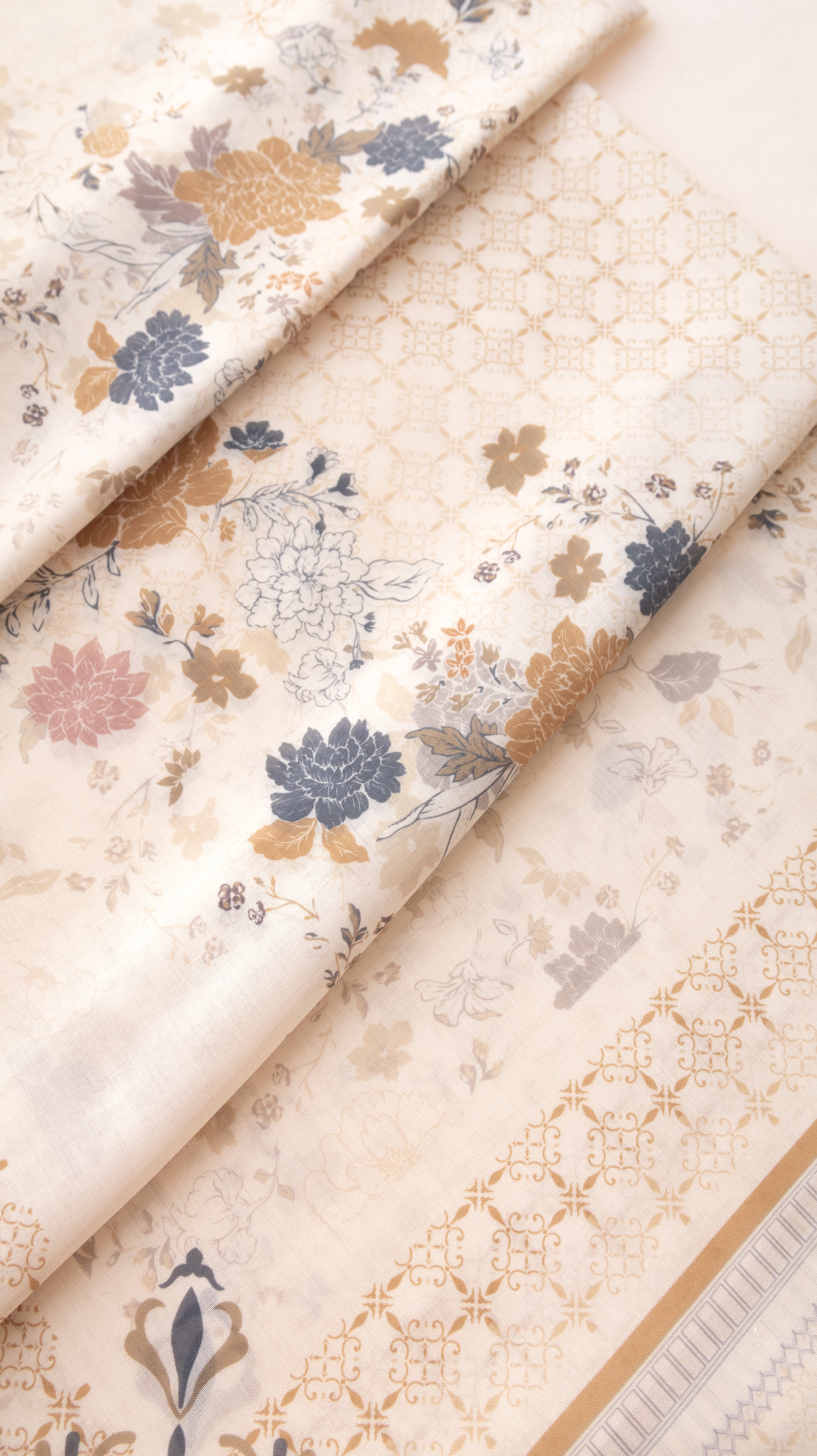 A folded piece of cream-colored fabric with a traditional floral print in muted blue, gold, and pink, featuring a matching gold geometric border.