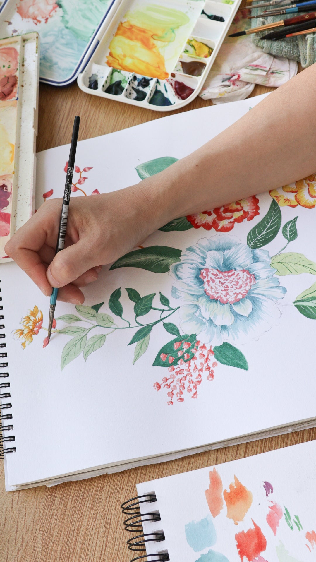 A close-up of a hand holding a paintbrush and painting a floral illustration in a spiral-bound sketchbook. Watercolor paints, brushes, and a palette are arranged on a wooden table.